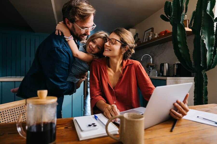 Parents and child smiling together in a kitchen while the mother works on her laptop.