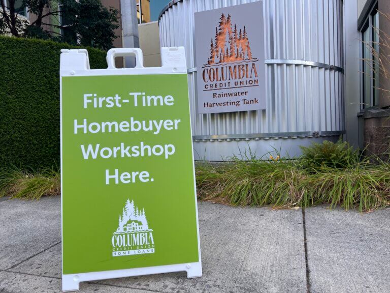 A green sign that reads "First-Time Homebuyer Workshop Here." sitting on the sidewalk in front of Columbia Credit Union's Operations Center (Ops).